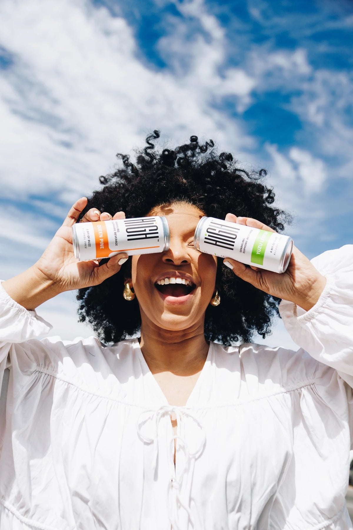 A smiling woman holding Queen Pine and Yuzu Lemon HIGH cans up to her eyes