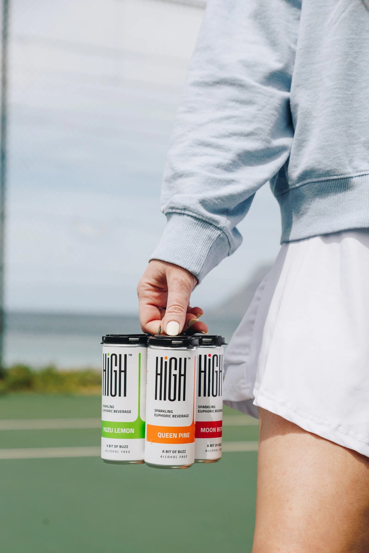 A person carrying a four-pack of HIGH cans beside a tennis court